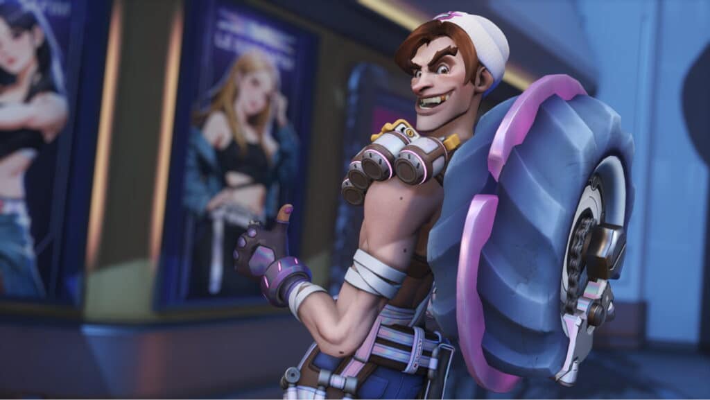 Junkrat from Overwatch 2 holds a large pink and blue tire-shaped shield, positioned against vibrant posters in a futuristic setting