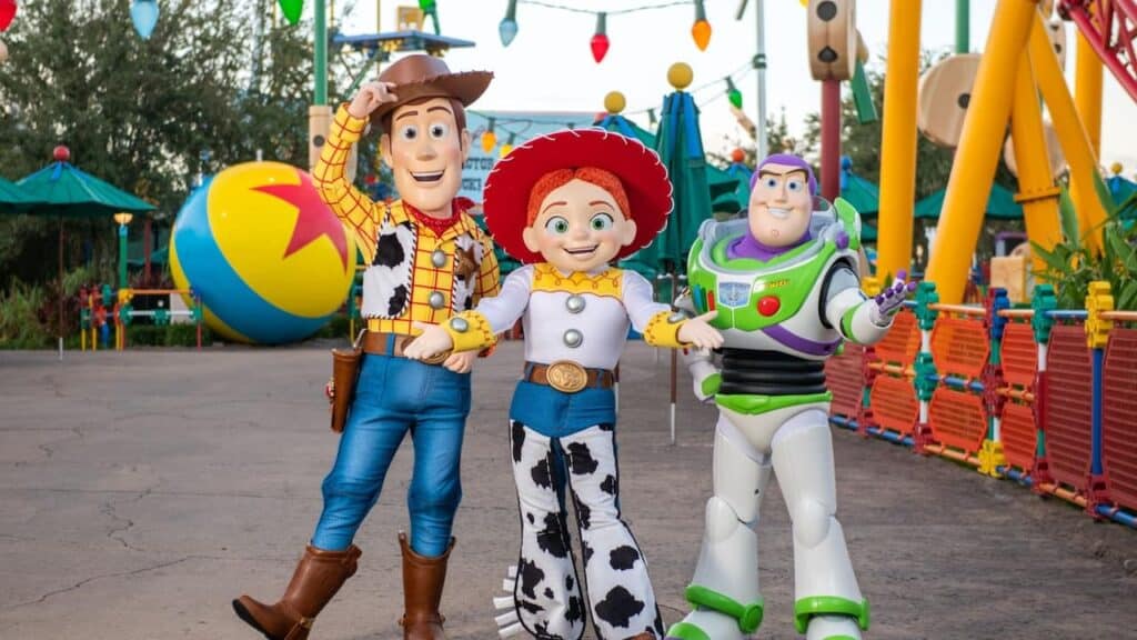 Toy Story characters in a theme park pose joyfully. From left: a cowboy, a cowgirl with red hair, and a space ranger. Bright, colorful setting with festive lights.