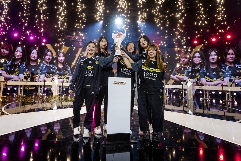 Five young women in esports jerseys proudly hold a trophy on stage, surrounded by celebratory lights and a large digital screen displaying their team image