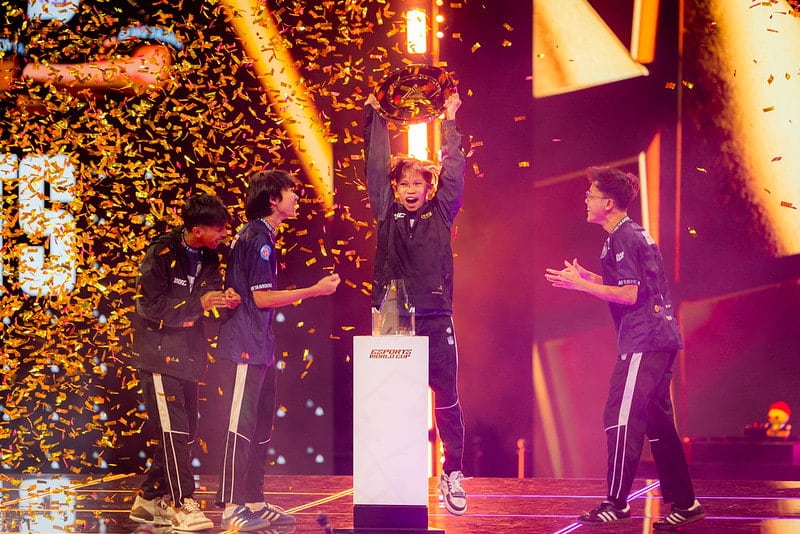 Four jubilant esports players in matching uniforms celebrate a victory on stage, one lifting a trophy amid golden confetti, under dramatic lighting