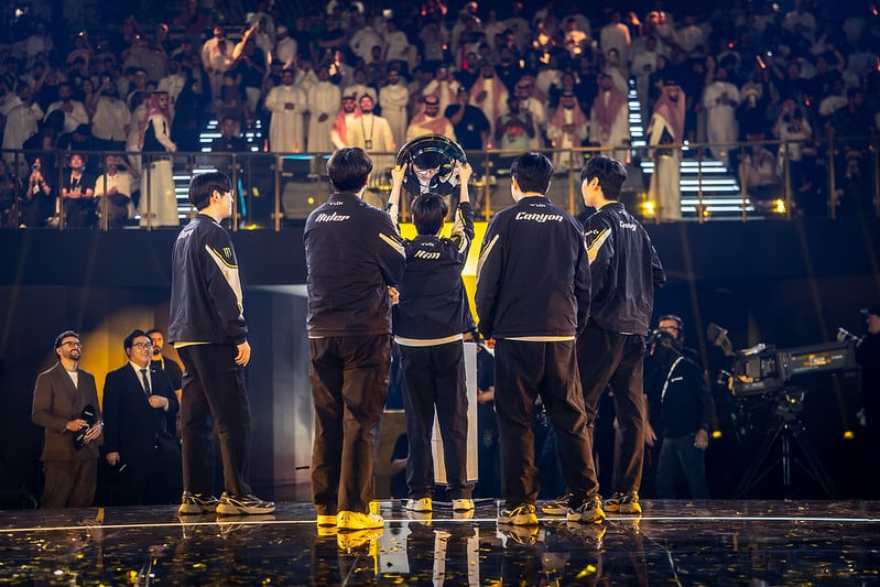Team lifting a trophy on stage under bright lights, watched by a cheering crowd