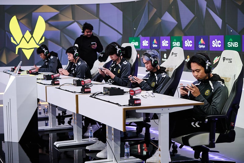 Five esports players sit in gaming chairs, focused on their phones during a competitive match. The atmosphere is intense and professional, with a large logo and brand banners in the background
