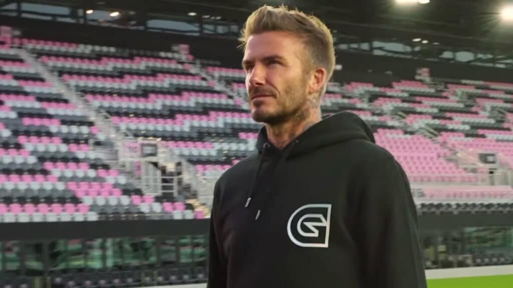 David Beckham wearing a black hoodie, stands confidently in an empty stadium with pink and black seats
