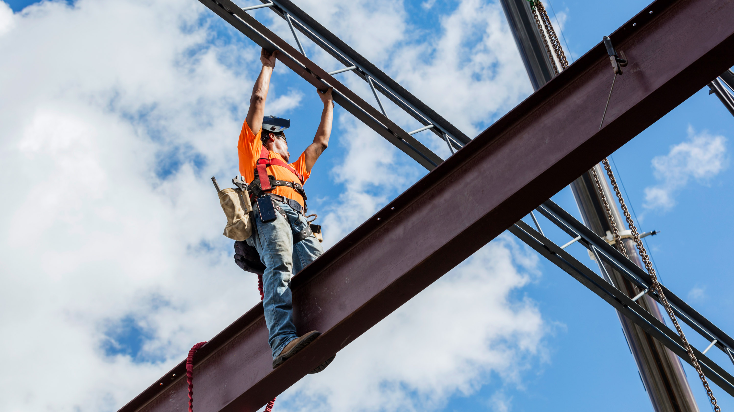 Steel worker in safety gear standing high up on a girder and reaching up overhead for a roof joist