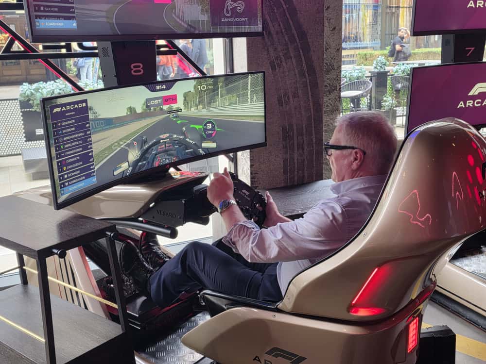 Richard Parker in a white shirt and sunglasses plays a Formula 1 racing simulation game in an arcade setting